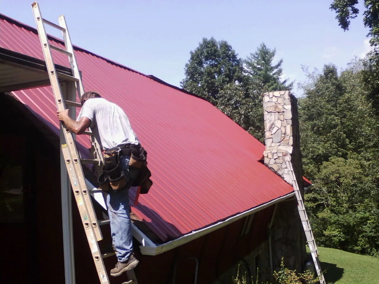 Roofer on a ladder beside a red metal roof and stone chimney — Soffit & Fascia Repair by Roofing Brothers in Hunt Valley